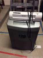 Front view of a Fellowes paper shredder with a Scotch laminator and bag sealer placed on top, showing power cord and general condition.