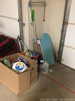 Wide view of cleaning supplies with mop bucket, mini ironing board, and box of assorted cleaning items near a garage door.