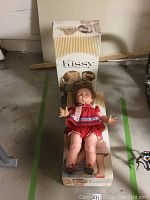 Front view of one vintage Kissy doll in red dress lying on top of its original box showing facial features and shoes.