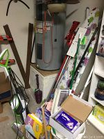 Photo showing Bissell vacuum cleaner, mop bucket, plunger, Kleenex box, assorted cleaning supplies, and ironing board in corner near water heater.