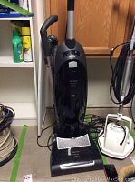 Full view of black upright Miele vacuum with hose, handle, and power cord in background.