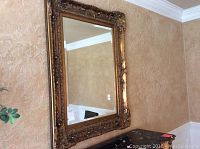 Full view of the rectangular mirror mounted on a textured tan wall, showing the elaborate gold frame detailing.