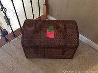 Closed brown wicker trunk with domed lid and front buckle straps on hardwood floor.