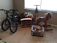 Photo showing two black kids' bikes, a variety of stuffed animals in a bin, a wooden rocking horse, and a wooden organizer with small toy cars. A Pogoroo scooter stick stands near the window.