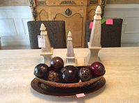 Seven ceramic ornamental balls arranged in a bronze hammered oval metal dish on a table with three light beige stone obelisk-shaped ornaments behind.