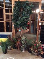 Photo showing one large faux tree with dark green leaves and brown trunk, surrounded by eight faux plants and floral arrangements of various colors and sizes placed on the floor.