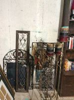 Photo showing five pieces of ornate metal wall art leaning against a wall alongside the plant stand and three tall candle stands with multi-colored candles on top.
