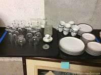 View of assorted wine glasses, tea cups, saucers, plates, and a dessert dish arranged on a black table against a bare concrete wall.