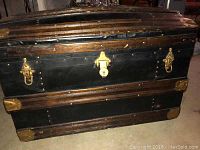 Front view of large black metal and wood trunk with brass hardware and locking mechanism, showing aged wear and scratches