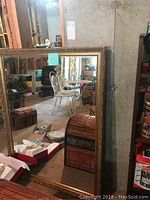 Large gold framed mirror leaning against a wall in basement environment, showing reflective surface and ornate detailed frame.