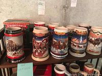 View of 7 Budweiser beer steins arranged in a row on a shelf with 3 visible Budweiser boxes behind them. Decorative Christmas and winter holiday scenes with Clydesdales pulling wagons on the steins.