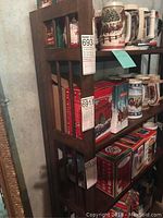 Side view of folding wood shelf with tags attached, showing vertical slats and dark wood finish. Shelves hold decorative steins and boxes, which are not included.