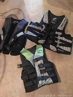 Three life jackets in shades of black, grey and green laid out on floor showing front buckles and straps for secure fit.