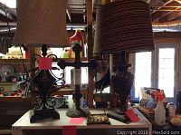 Two decorative lamps and decorative iron cross shown on table with background of workshop or garage. Lamps have fabric shades; one beige pleated, one reddish striped. Iron cross is rustic metal with round base.