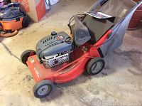 Front-side view of the Scotts lawn mower showing red deck, black engine casing, fabric grass catcher, and wheels.