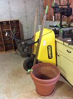 Yellow Stanley poly tub wheelbarrow with wooden handles and flat tire, positioned next to a large clay pot in a basement or garage setting.