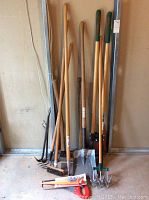 Photo showing multiple wooden handled yard tools including shovel, pick axe, post hole digger, aerator, and hoe, leaning against a wall.