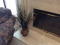 Faux plant in brown ceramic pot with long green and beige stalks beside fireplace and floral couch.