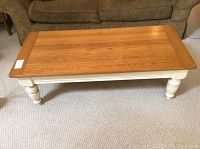 Front view of rectangular coffee table showing light oak wood top and cream painted turned legs on carpeted floor.