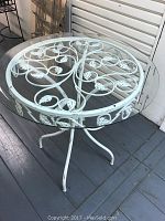 Top angle of the bistro table showing the glass top and decorative white metal scroll and leaf pattern base.