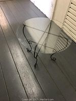 Round glass coffee or side table with a clear removable glass top and black folding metal leg stand, shown from a side angle on a wood floor.