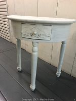 Front view of small white semi-circular table with single decorative drawer and four turned legs.