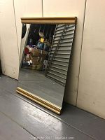 Front view of rectangular mirror showing reflective surface and gold trim on top and bottom edges, placed leaning against a wall.