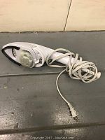 Image of handheld clothes steamer lying flat on a floor, with visible steam plate, textured brush attachment, and power cord with plug.