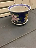 Photo of a round ceramic flower pot with a blue background and pink and yellow floral patterns. White rim with blue decorative band. Pot sits on matching ceramic saucer. Surface shows minor signs of use and some dirt inside.