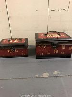 Two closed rectangular Asian-style boxes with red and black finish and gold calligraphy on the exterior against a plain background.