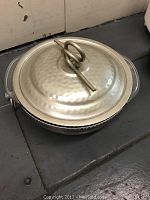 Front angle showing hammered tin casserole dish with lid and loop handle on top, placed on a floor surface.
