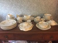 Photo of full china set arranged on wooden dresser including plates, bowls, cups, covered serving dishes.