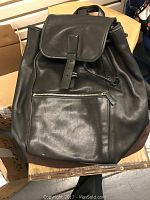 Front view of black leather backpack with flap buckle pulled down and front zipper pocket visible.