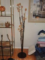 Full view of the large floor-standing bud vase on a wood floor near a shelving unit and pile of fabric