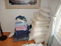 Photo showing a stack of white and cream colored pillows of various sizes stacked near window with some folded linens and towels in the foreground.