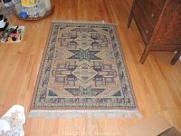 Rectangular woven area rug with traditional geometric patterns in tan, blue, rust colors, fringed edges, placed on wood floor.