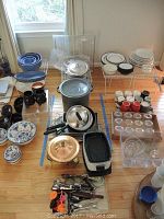 Overview of lot contents with pots, pans, racks, serving dishes, plates, and kitchenware arranged on wooden floor.