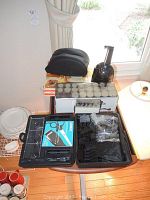 Photo showing electric hair curlers in plastic case, Braun shaver charger, black zippered carrying case, and wooden table next to window