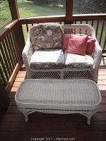 Wicker loveseat with floral seat cushions and pink pillows situated on wooden deck.