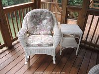 Front view of white wicker rocking chair with floral cushions and white wicker side table on wooden deck.