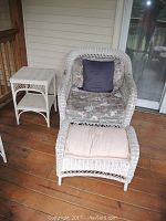 Set of white wicker furniture on wooden porch: chair with floral cushions and a dark small cushion, ottoman with cushion, and side table with two shelves