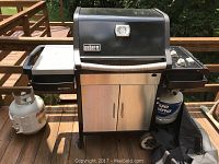 Weber Spirit E-310 gas grill shown from front with propane tank on deck. Stainless steel storage cabinet and three burner control knobs visible.