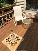 Two glass and metal side tables, white web chair, and wrought iron umbrella stand on outdoor wood deck.