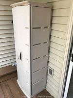 Overall view of a tall vertical plastic outdoor storage cabinet in off-white color with a single door and molded handle, showing signs of soil and wear.