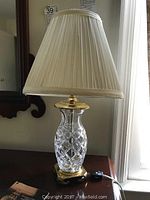 Photo showing full lamp on table with pleated cream fabric shade and brass base, lamp plugged in with switch on cord.