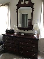 Full view of the mahogany dresser with brass handles and separate ornate mirror mounted above, placed between two windows.