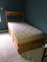 Twin wooden bed against a teal wall with mattress, showing front and side view of closed drawers and door on the bed frame.