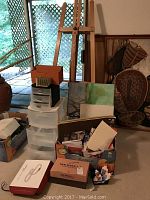 Wooden easel leaning against wall next to plastic storage drawer units. Cardboard boxes contain art supplies and papers. Several canvases and paintings behind the drawers.