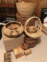 Photo of various woven baskets including a large basket with handle on top, a round basket bowl with dark pattern, a storage cube basket, and a decorative painted gourd placed on wooden boxes in front.