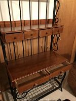 Full view of bakers rack showing wooden top drawers, middle wooden surface, and bottom metal wire shelf with decorative scroll metalwork.
